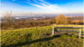 Eine Holzbank auf einem grasbewachsenen Hügel mit Blick auf eine Stadtlandschaft im Hintergrund unter einem blauen Himmel mit weißen Wolken.
